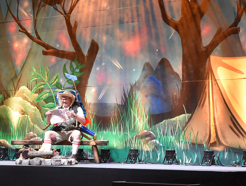 A person in a camping costume sits on a bench reading a map on a stage with a colorful forest and tent backdrop, illuminated by stage lights.