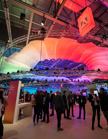 People gather at an indoor tech event beneath a large, colorful, wavy canopy with dramatic lighting. An Adobe sign hangs overhead and display booths are visible around the space.
