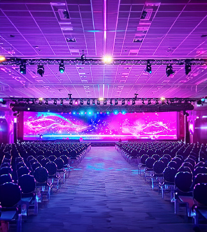 A large, empty event hall with rows of chairs facing a brightly lit stage. The stage backdrop displays vibrant pink and blue swirling lights under a high ceiling with spotlights.