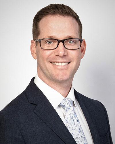 A man wearing glasses and a dark suit with a white shirt and patterned tie smiles at the camera against a plain light background.