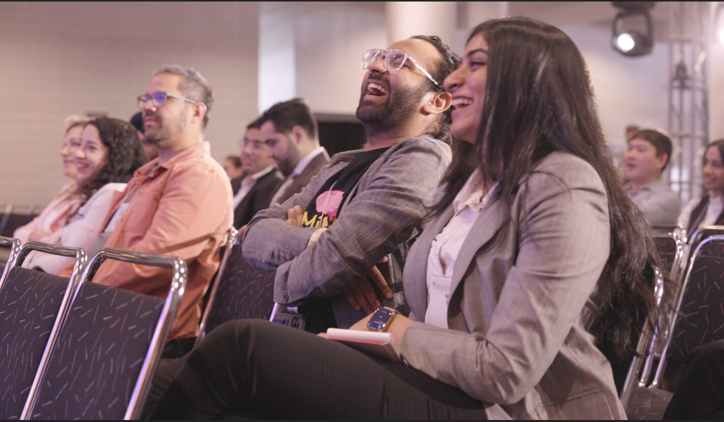 A group of people sit in rows of chairs at an indoor event, smiling and laughing as they watch something off-camera. The atmosphere appears relaxed and cheerful.