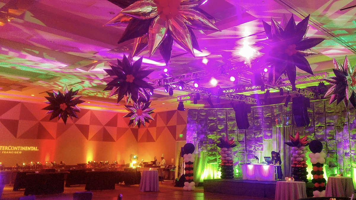 A ballroom decorated with large star-shaped balloons, colorful lighting in purple and green, and a DJ booth on stage surrounded by balloon columns; tables are arranged around the room.