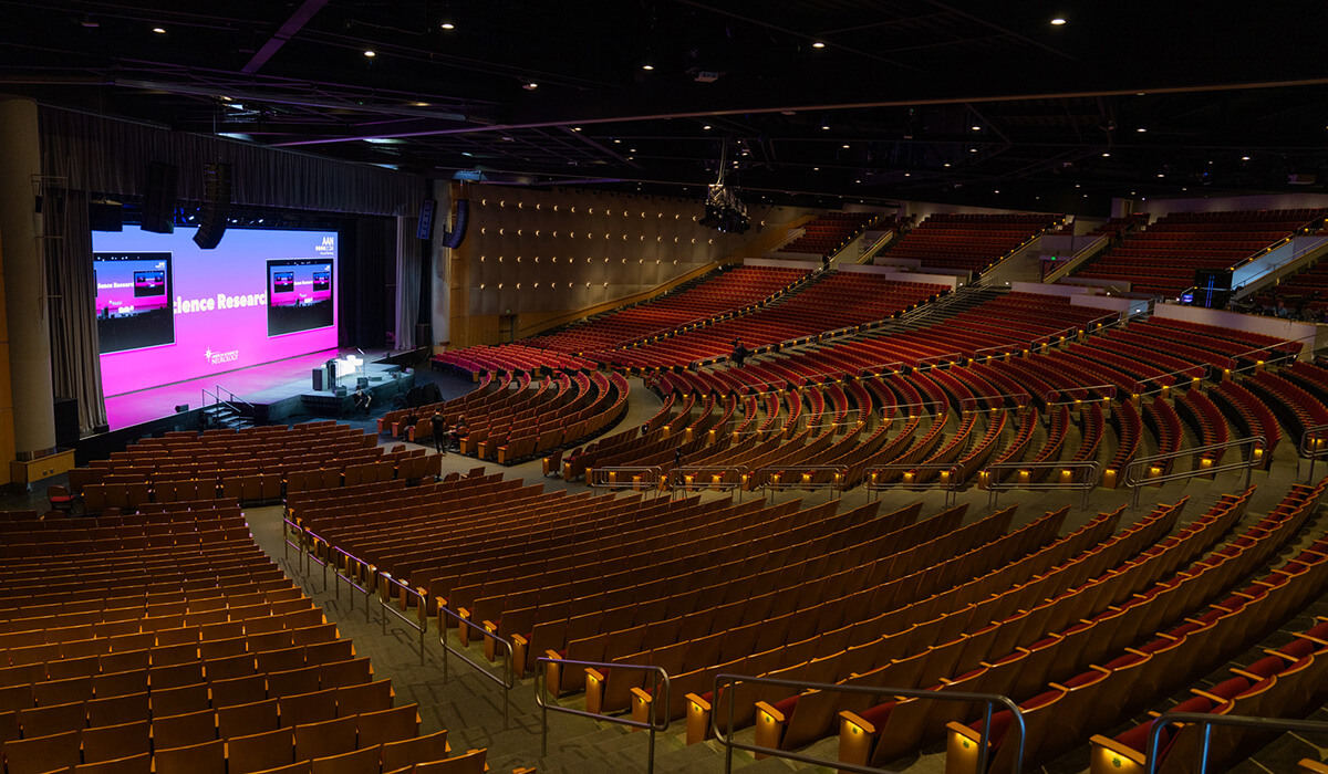 Large, empty auditorium with rows of brown seats facing a brightly lit stage with a large screen displaying a pink background and text. Stage setup includes podiums and presentation equipment. Ceiling lights illuminate the space.