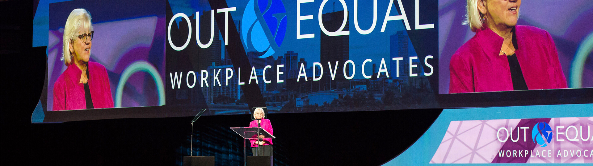 A person in a bright pink jacket speaks at a podium on stage in front of a large screen displaying OUT & EQUAL WORKPLACE ADVOCATES and their magnified image.