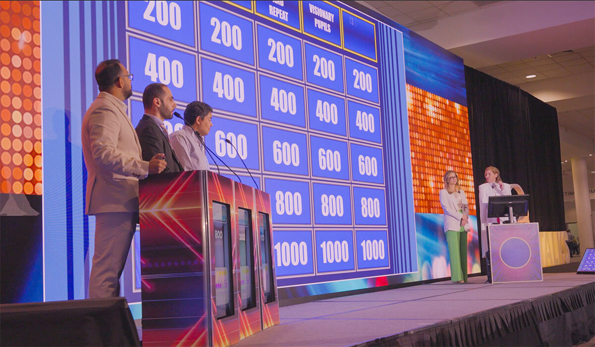 Four people stand at podiums playing a Jeopardy-style game, facing a large screen displaying the game board with point values. Two hosts are at a computer on the right side of the image.