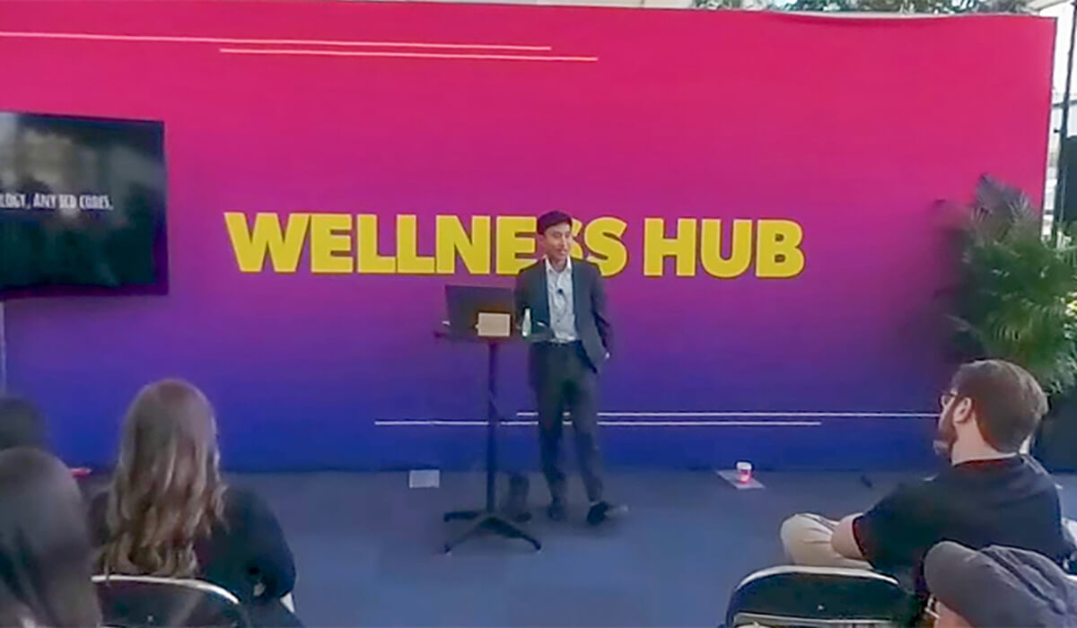A man in a suit stands at a podium giving a presentation to a seated audience. Behind him is a large, colorful sign that reads WELLNESS HUB. A TV screen and potted plants are visible on either side.