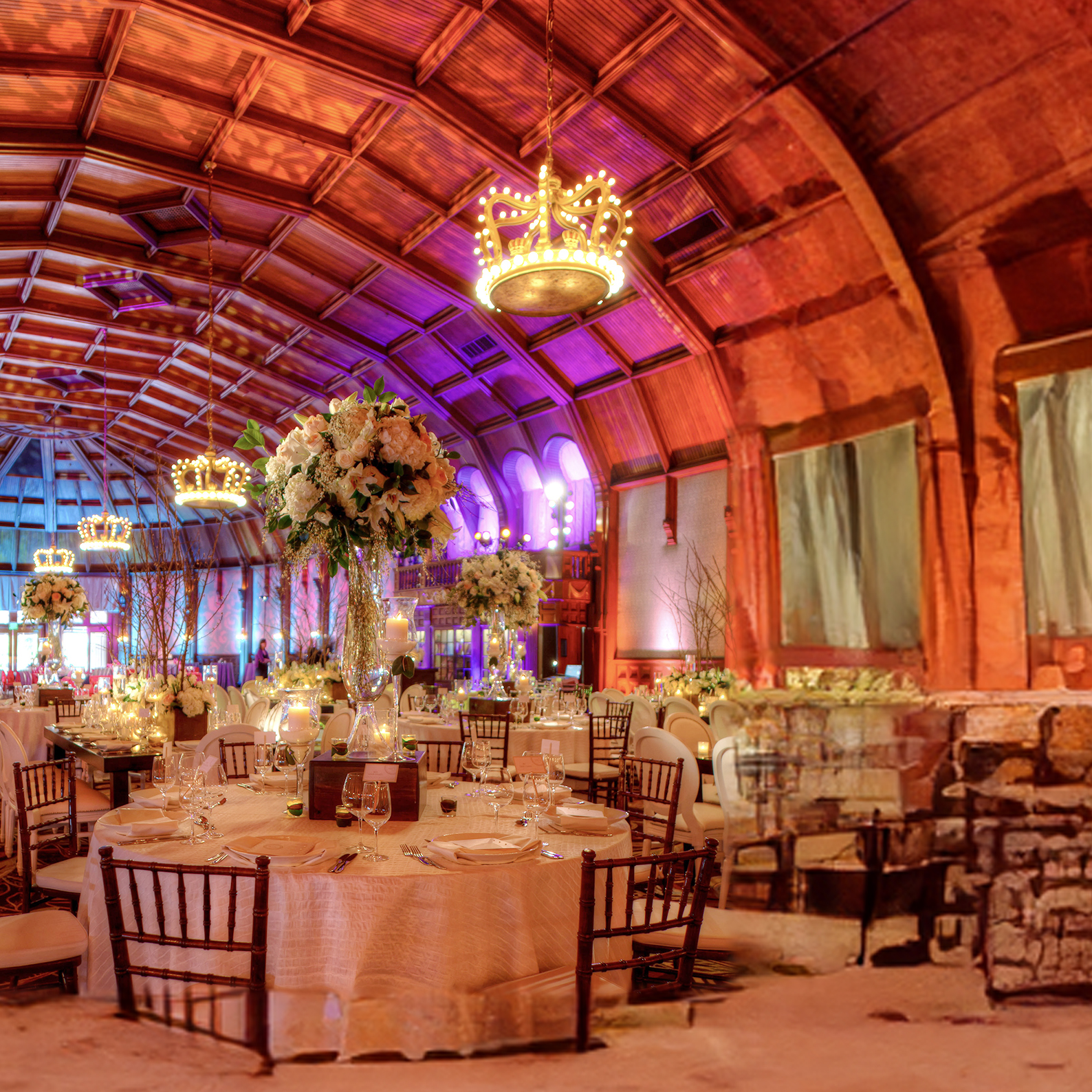 A grand banquet hall with a high arched ceiling, chandeliers, round tables set with elegant linens, floral centerpieces, and wooden chairs, all illuminated by warm, colorful lighting.