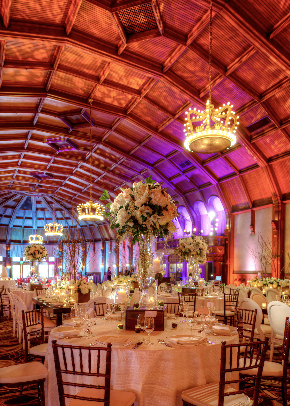 A lavish ballroom set for an event, with round tables dressed in white linens, tall floral centerpieces, elegant tableware, and wooden chairs. Warm lighting and grand chandeliers hang from a vaulted wooden ceiling.
