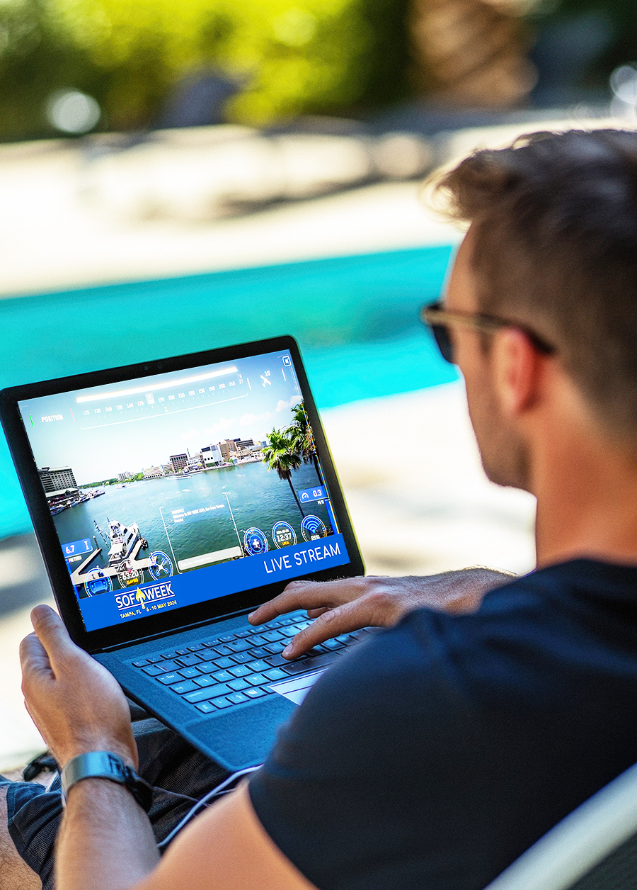 A man wearing sunglasses uses a laptop by a swimming pool, watching a live stream of a boat racing event on the screen. The background is bright and sunny, suggesting a relaxed outdoor setting.
