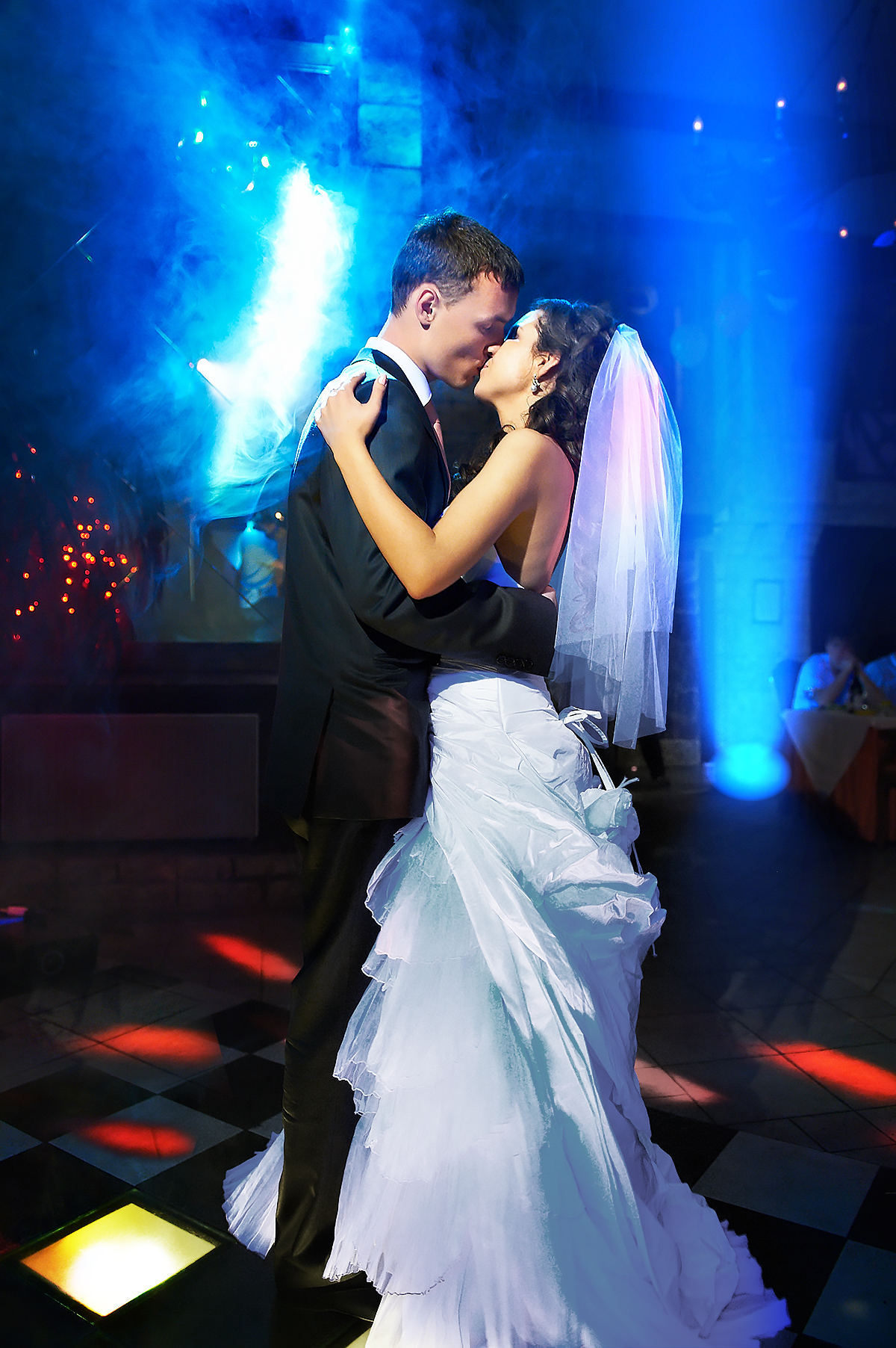 A bride and groom share a romantic first dance on a checkered floor, bathed in colorful stage lights and surrounded by blue smoke, creating a dreamy wedding atmosphere.
