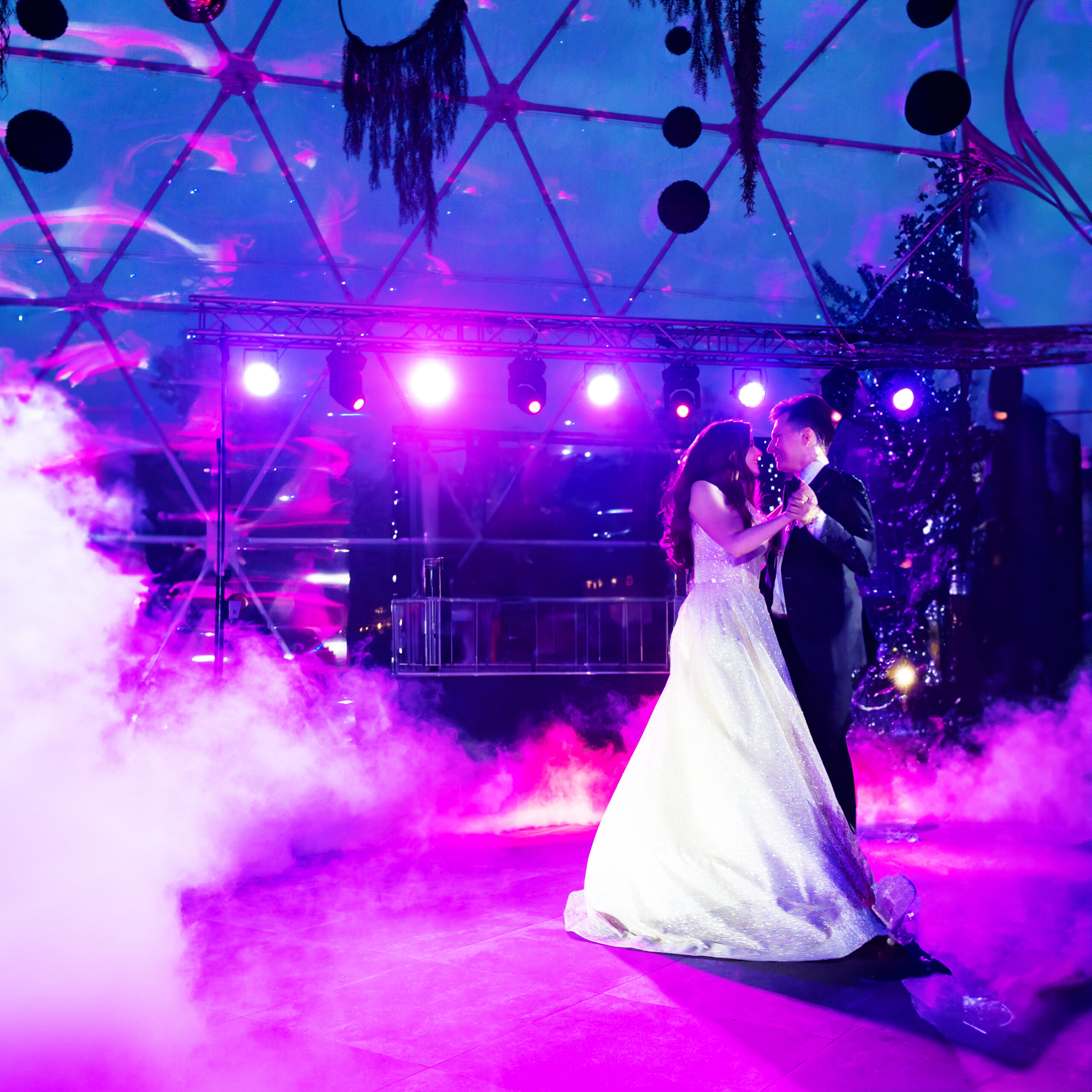 A bride and groom share their first dance under purple lighting, surrounded by fog, inside a modern, dome-shaped venue with a vibrant, dreamy atmosphere.