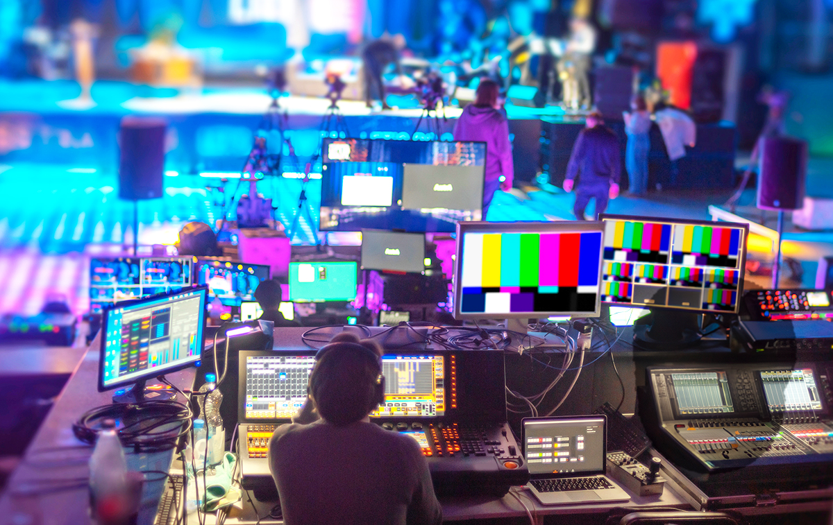 A person operates sound and video control boards in a studio filled with screens, computers, and technical equipment, as people prepare a brightly lit stage area in the background.