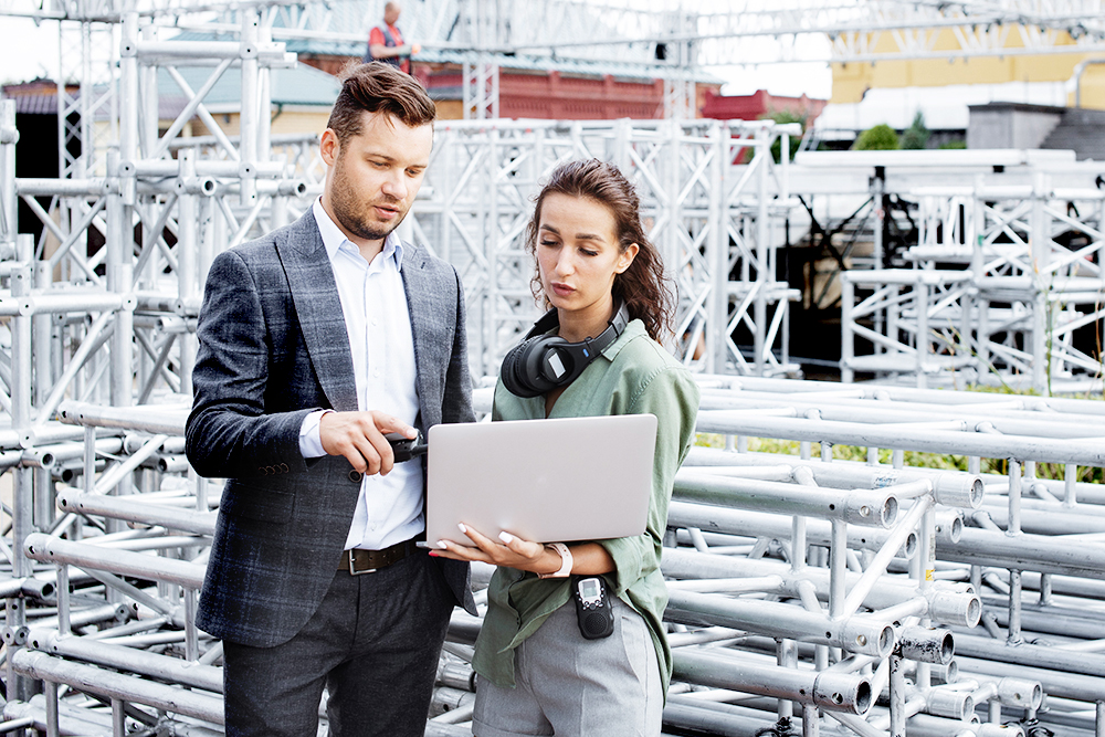 A man in a suit and a woman with headphones around her neck stand outdoors among metal scaffolding, discussing something while looking at a laptop the woman is holding.
