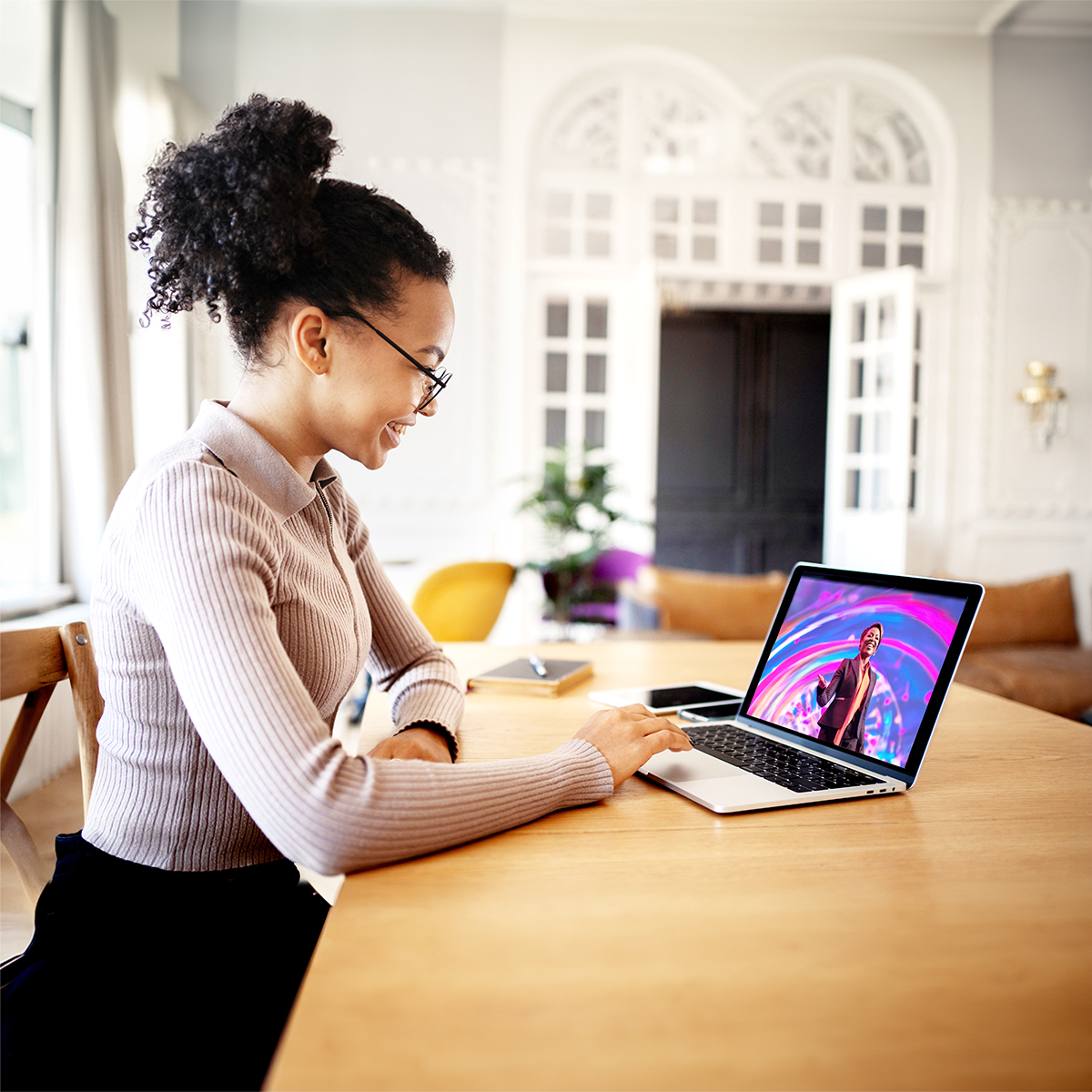 A woman sits at a wooden table in a bright room, smiling as she uses a laptop. On the laptop screen, a person appears on a colorful stage, suggesting a video call or virtual event.