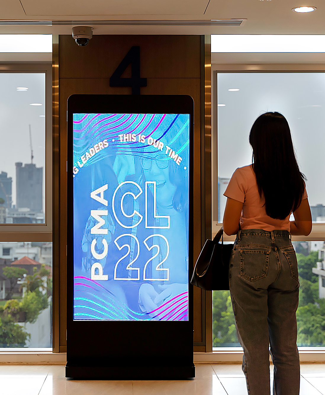 A woman stands in front of a digital sign displaying “PCMA CL22” and “LEADERS THIS IS OUR TIME” in a modern building with large windows overlooking a city skyline.