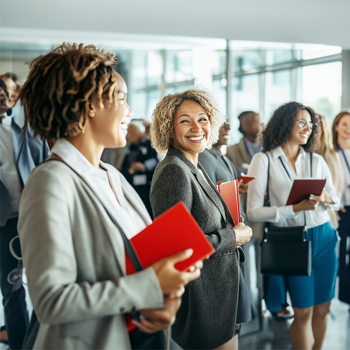 A group of professionally dressed people, some holding red folders, stand smiling and talking in a bright, modern office space.
