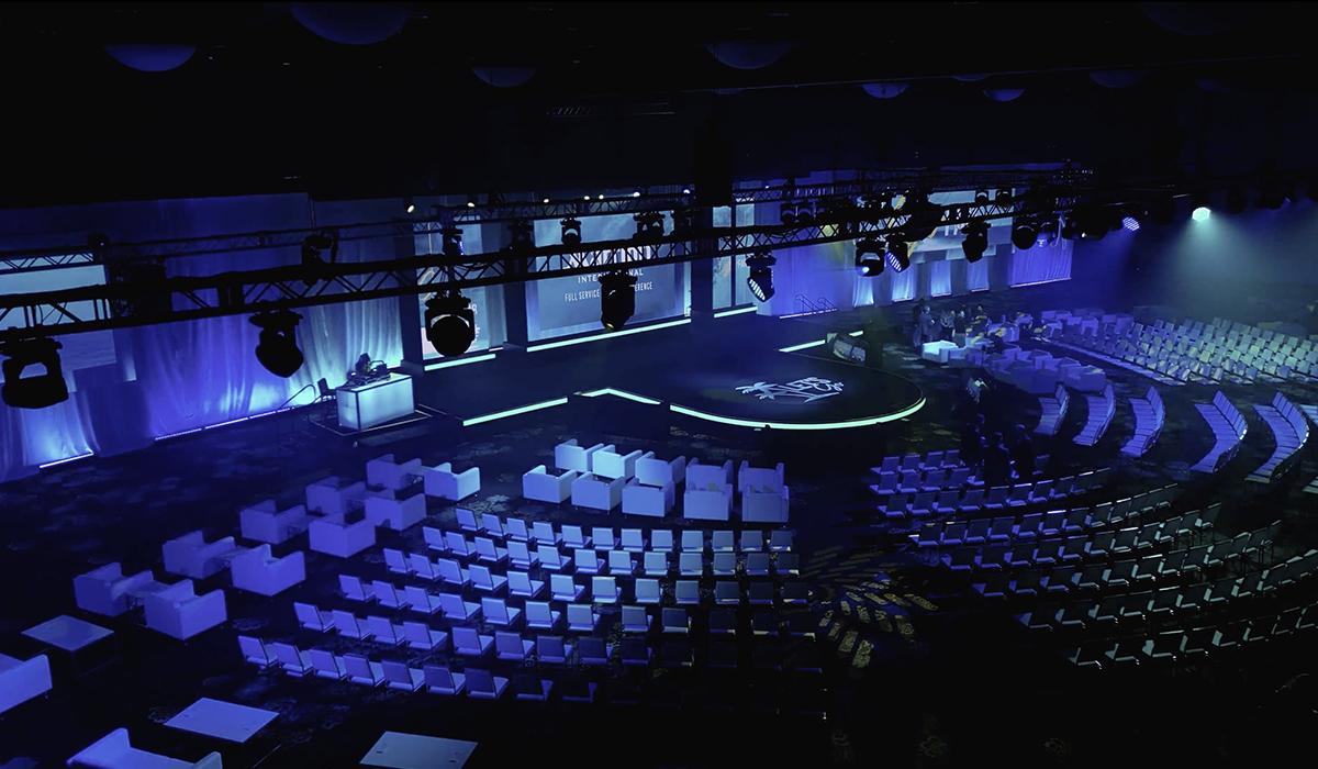 A large, dimly lit auditorium with rows of empty seats and a modern stage illuminated in blue and purple lighting, prepared for an event or conference.