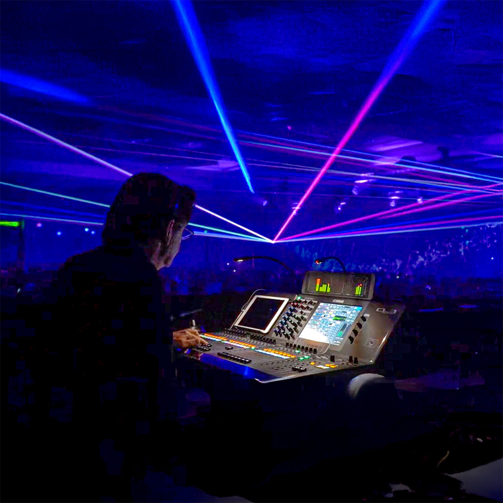 A sound engineer operates a mixing console at a concert, with colorful laser lights and a large crowd visible in the background.