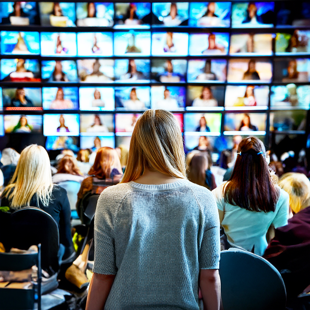 A group of people sit and stand facing a large wall of multiple television screens, each displaying different images, creating a busy and high-tech atmosphere.