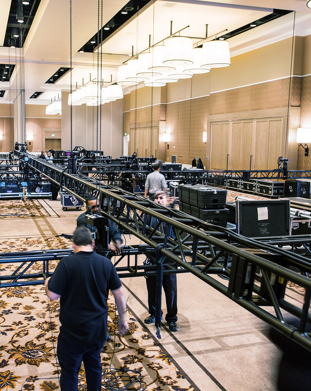 People assemble a large metal truss structure in a hotel ballroom with patterned carpet, large light fixtures, and equipment cases. The ceiling and lights are reflected in a mirrored wall.