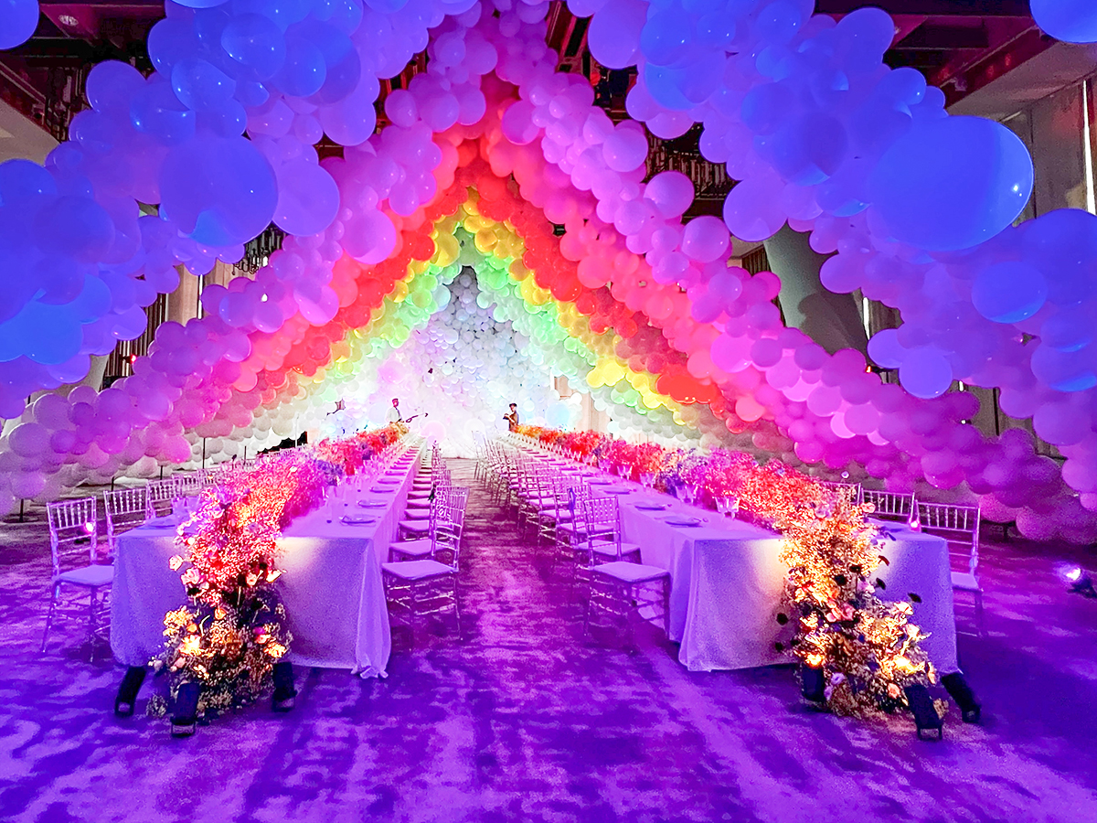 A vibrant event space features long banquet tables with white tablecloths and floral centerpieces under arches of colorful, illuminated balloons arranged in a rainbow pattern, creating a festive and whimsical atmosphere.