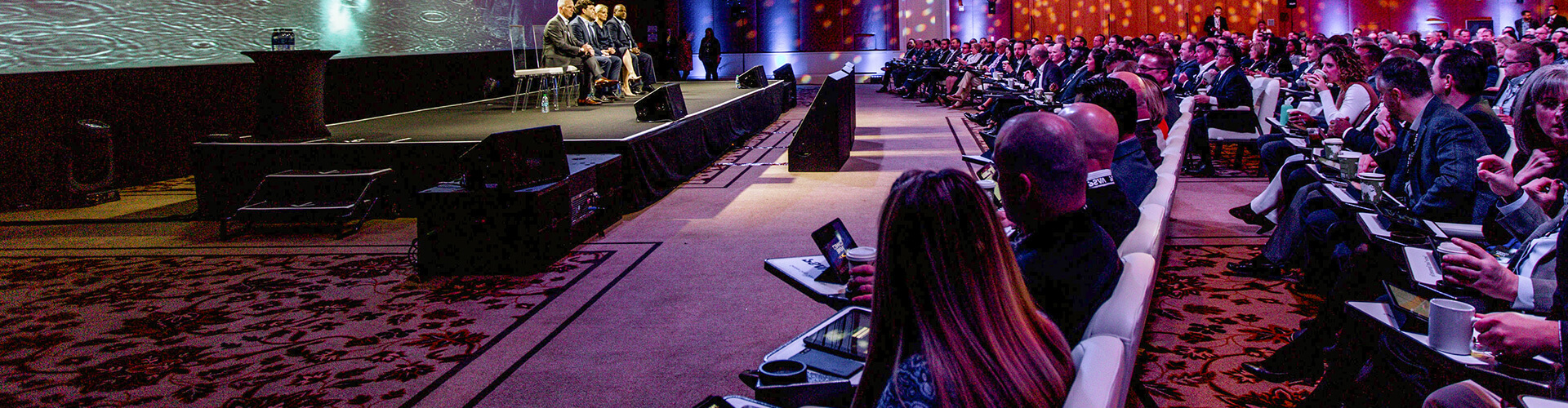 A large audience sits in rows facing a stage where several panelists are seated, speaking at a professional conference or business event. Many attendees hold notebooks or tablets. The room is brightly lit.