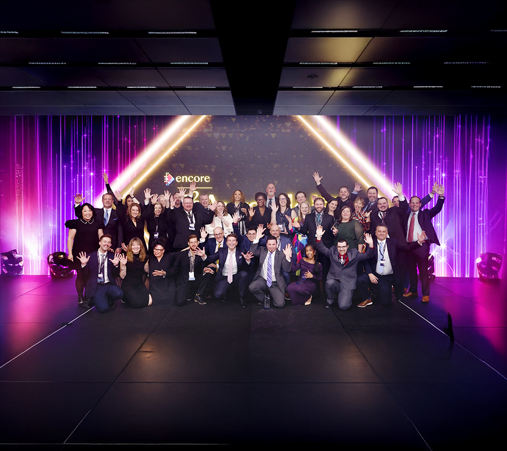 A large group of people in formal attire pose and smile on stage with raised hands in front of a glowing purple and gold backdrop featuring the encore logo.