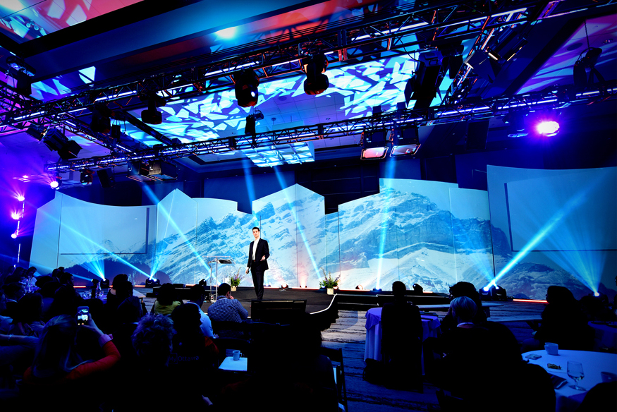 A speaker stands on a stage in front of an audience, with blue lights and a large backdrop displaying mountain scenery. The event space is filled with dramatic lighting and visual effects, creating an engaging atmosphere.