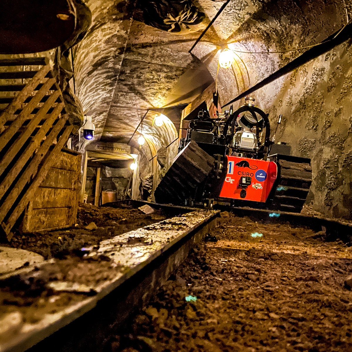 A small, red, tracked robot navigates a dimly lit, rough underground tunnel lined with dirt, rocks, and wooden supports, with lights hanging from the ceiling.