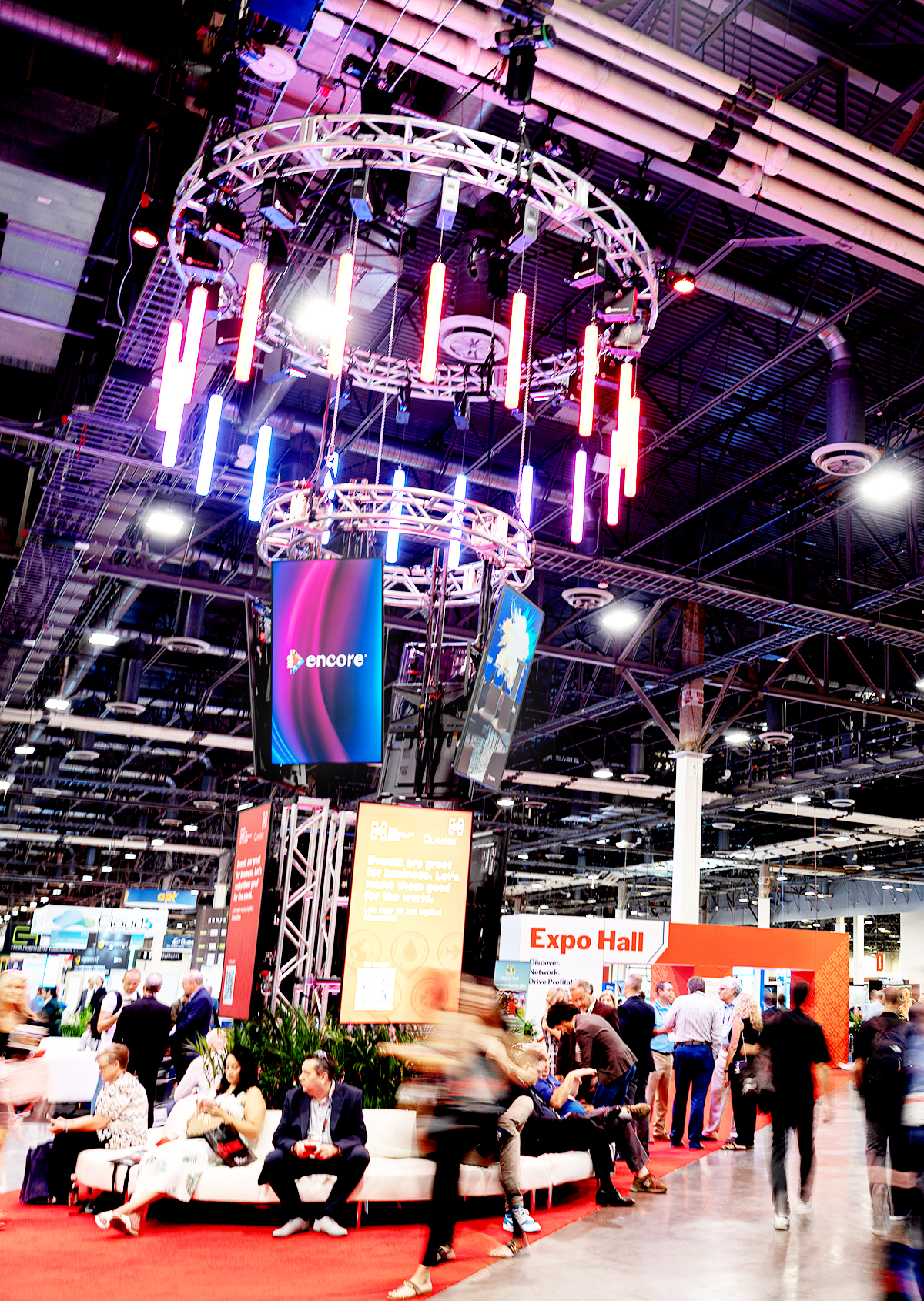 A busy convention center with people walking and sitting near a tall lighting display featuring colorful LED lights and screens. In the background, a red Expo Hall sign is visible.