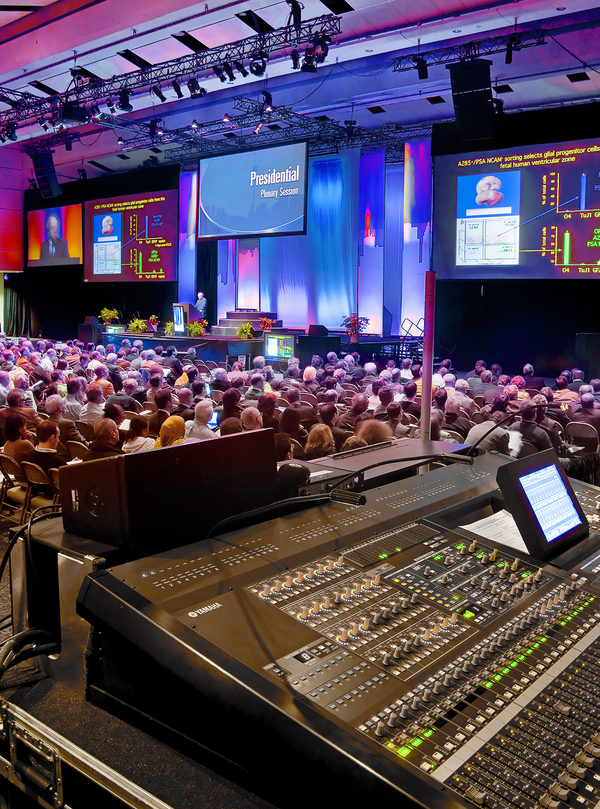 A large conference hall filled with people attending a presentation, featuring colorful stage lights, projection screens displaying slides, and an audio mixing console in the foreground.