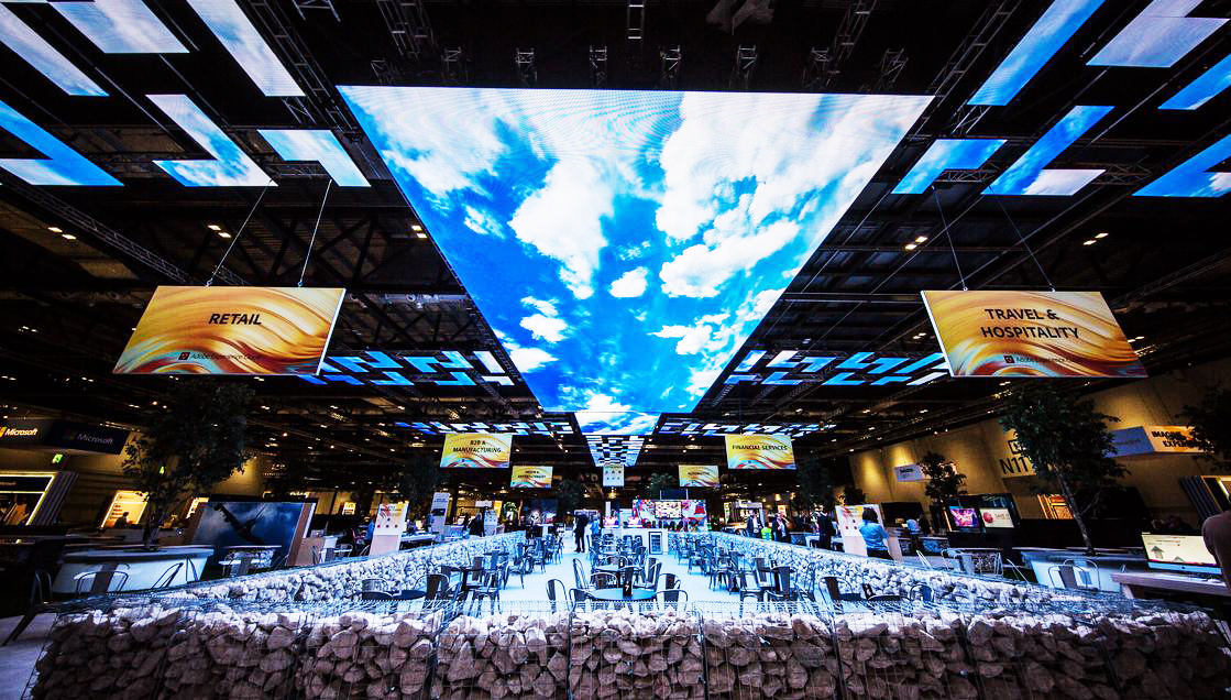 A large indoor exhibition hall features a dramatic ceiling display of LED screens showing a blue sky with clouds above tables and chairs. Hanging signs read Retail and Travel & Hospitality.