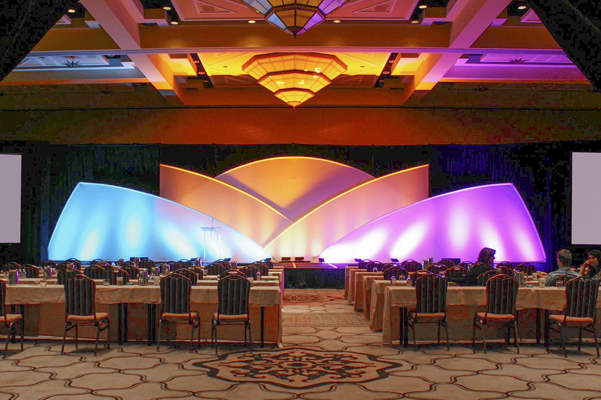 A conference room with a stage featuring colorful, curved light displays in purple, yellow, and orange. Several rows of tables and chairs face the stage, with a few people seated.