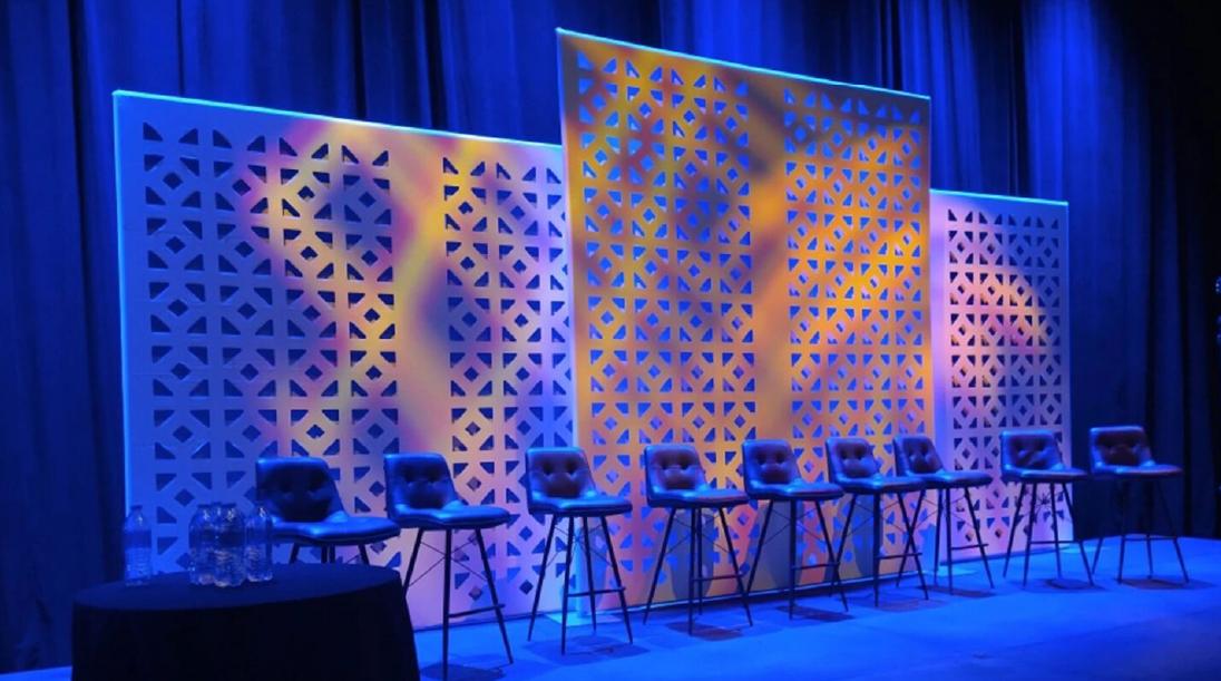 A stage with seven empty chairs in front of decorative panels, illuminated by blue and purple lights. A small round table with bottles and glasses is on the left side of the stage.