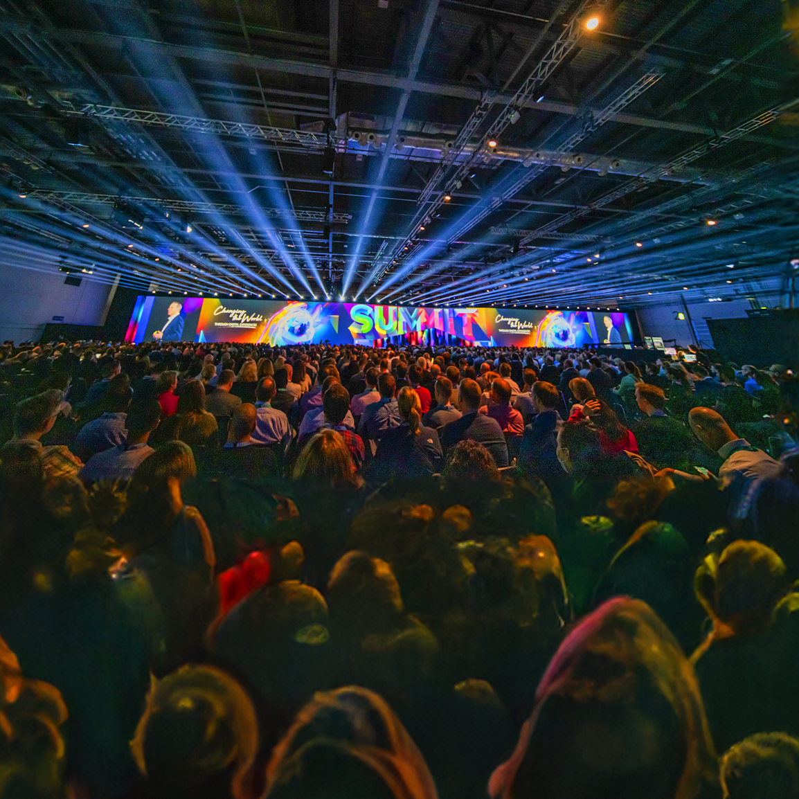 A large crowd sits in a dimly lit auditorium facing a brightly lit stage with colorful lights and the word SUMMIT displayed prominently on a screen, creating a vibrant atmosphere.