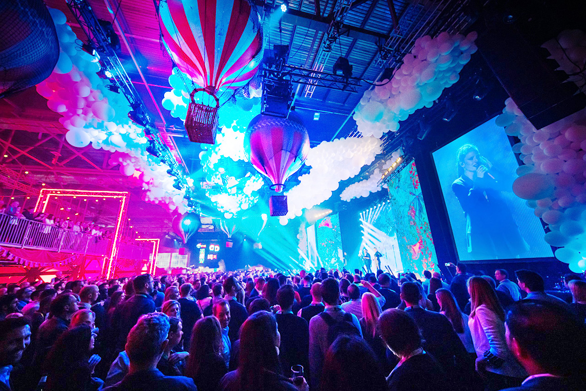 A lively crowd watches a performer on stage at a vibrant event with colorful lights, hot air balloon decorations, hanging white balloons, and a large screen displaying the singer.