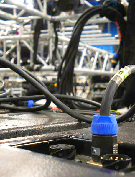 Close-up of audio and power cables connected to stage equipment, with blurred metal trusses and additional cables in the background, suggesting a live event or concert setup.