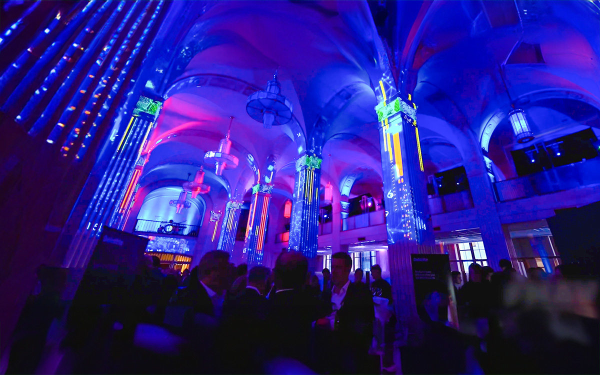 높은 기둥과 샹들리에를 비추는 생생한 파란색, 보라색, 분홍색 조명이 비추는 아치형 천장이 있는 웅장한 홀에서 한 무리의 사람들이 실내 이벤트에서 어울리고 있습니다.