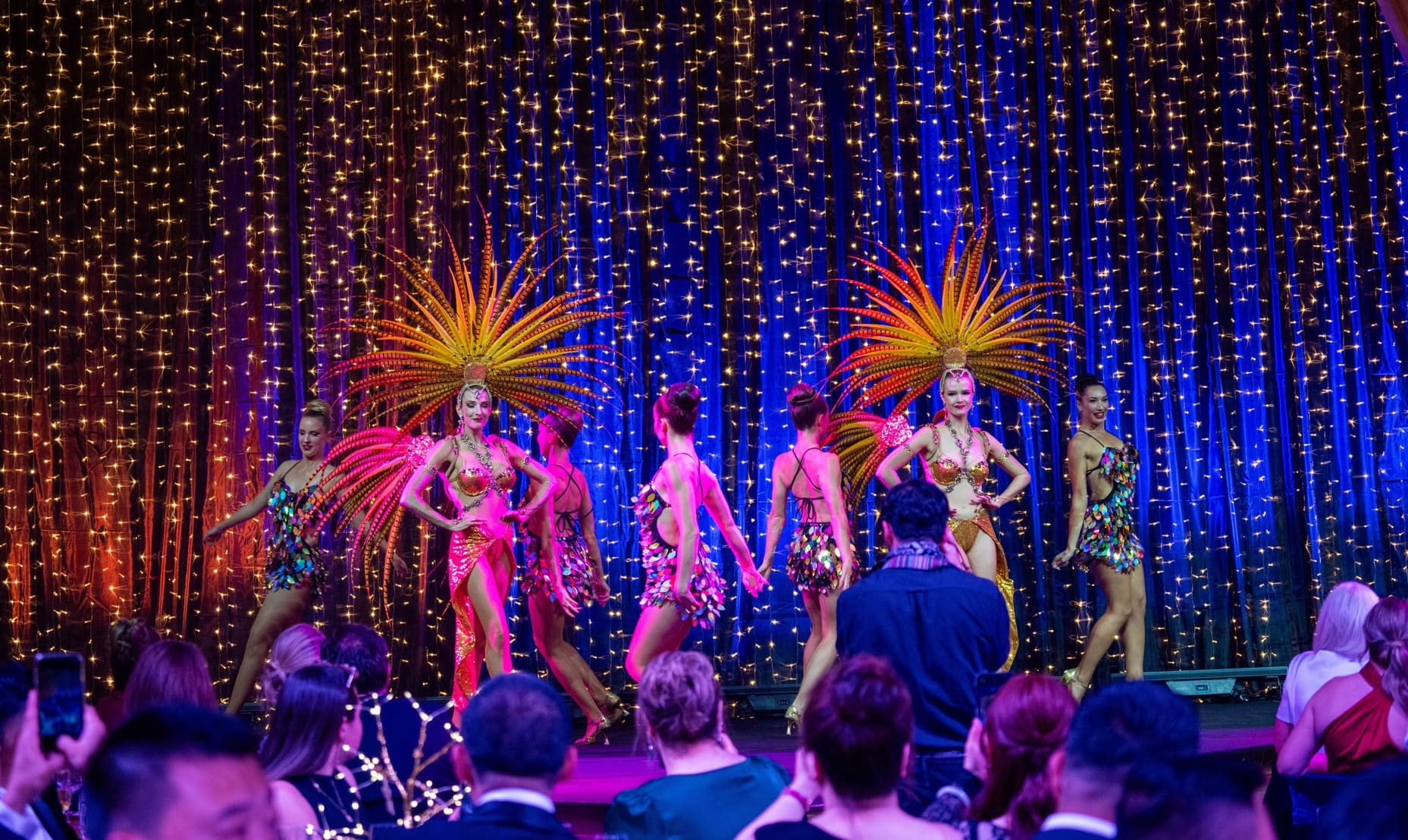 A group of female dancers in colorful costumes with large feathered headpieces perform on stage, with a sparkling curtain of lights as the backdrop, while an audience watches and takes photos.