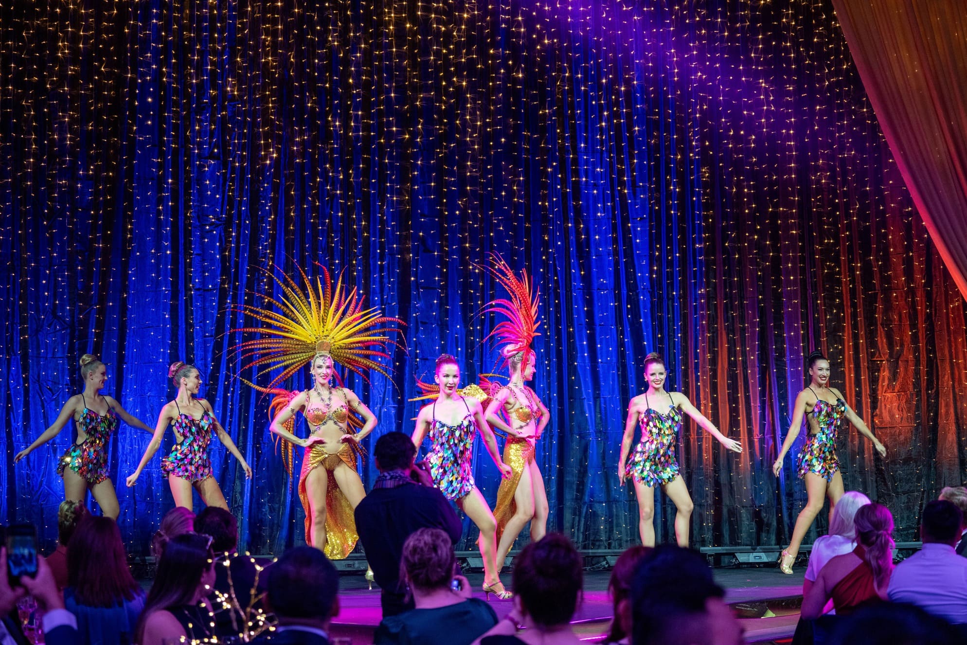 A group of female dancers in colorful, sparkling costumes and large feathered headdresses perform on stage under vibrant purple and blue lighting, while an audience watches.