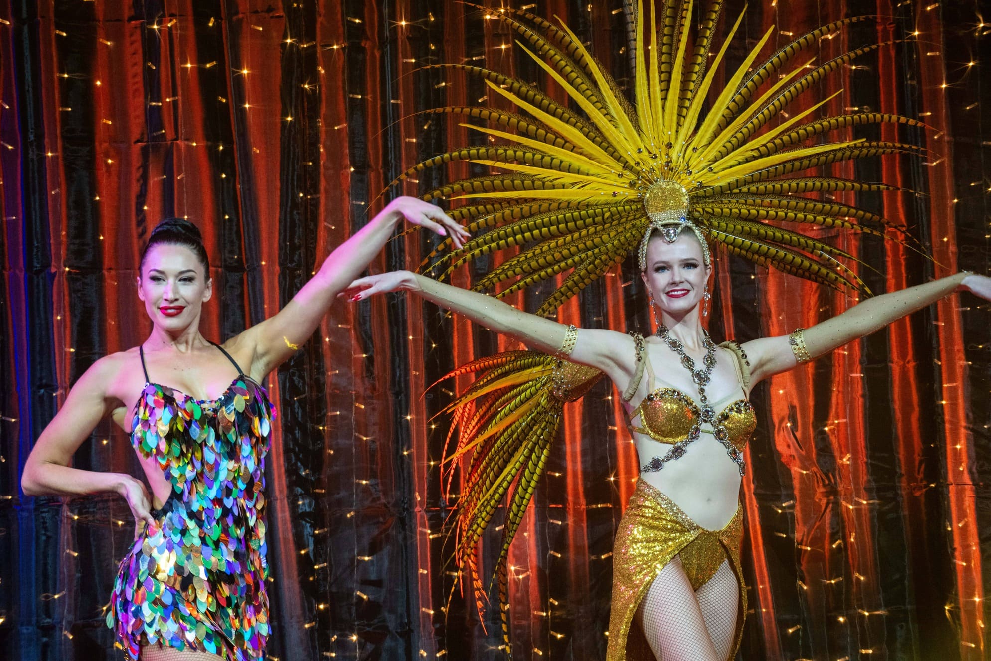 Two women pose on stage in front of sparkling lights; one wears a colorful, sequin dress, and the other wears a gold costume with a large, feathered headdress, both smiling with arms gracefully extended.