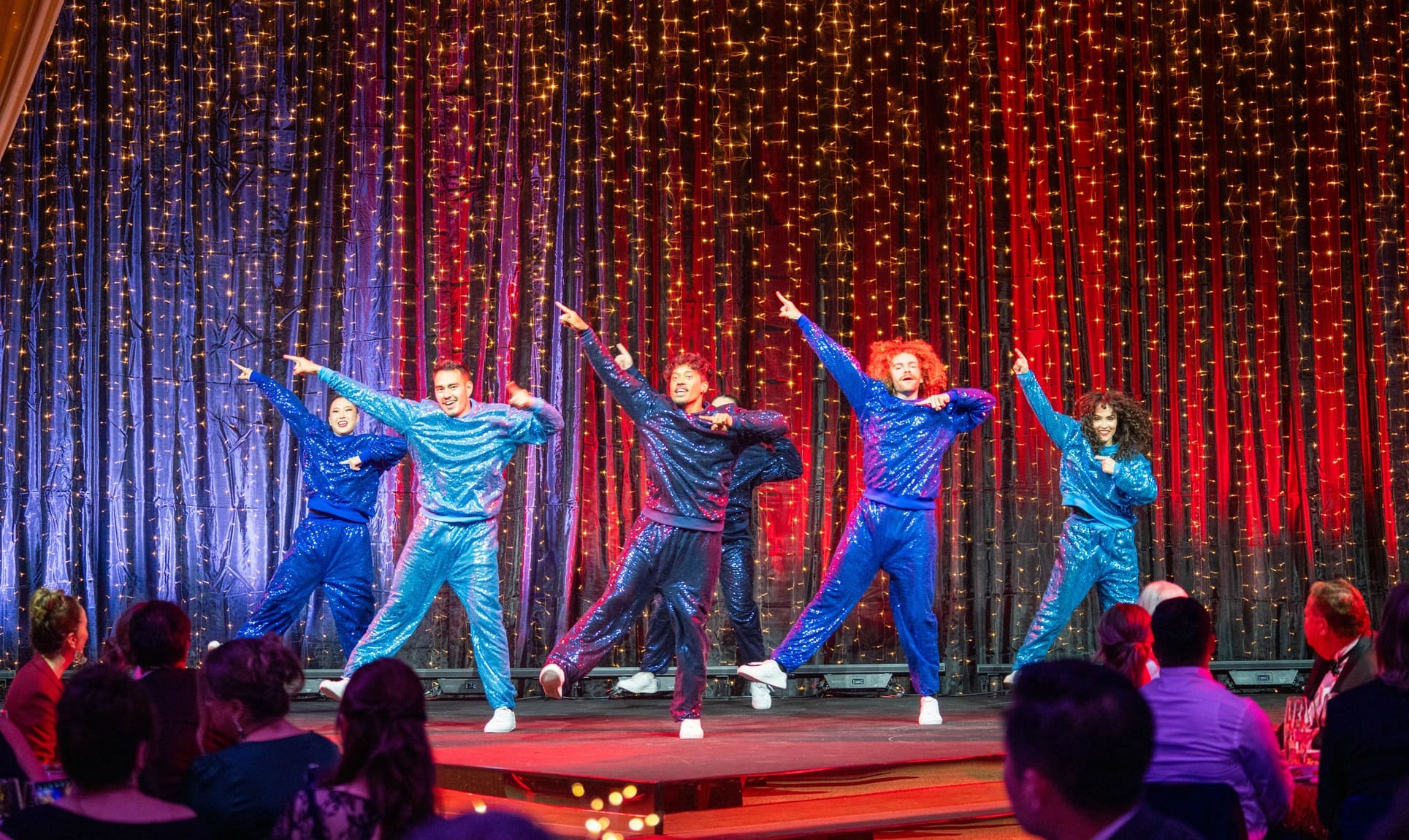 Five performers in matching blue and glittery jumpsuits dance enthusiastically on a stage with red and gold curtain lights, while an audience watches from tables in the foreground.