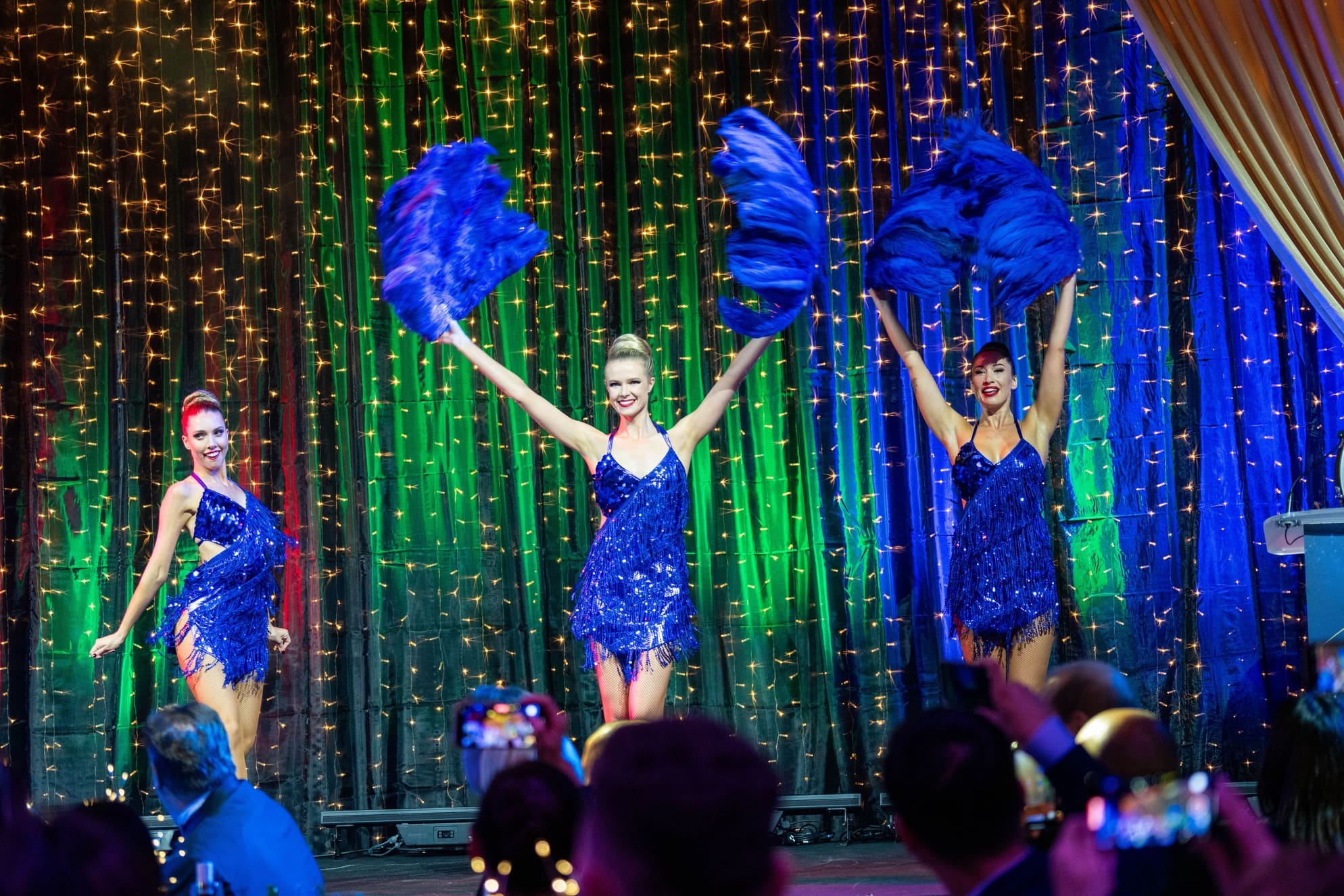 Three dancers in sparkling blue dresses perform on stage with blue feathered fans, smiling under colorful lights and a backdrop of hanging string lights, while an audience watches.