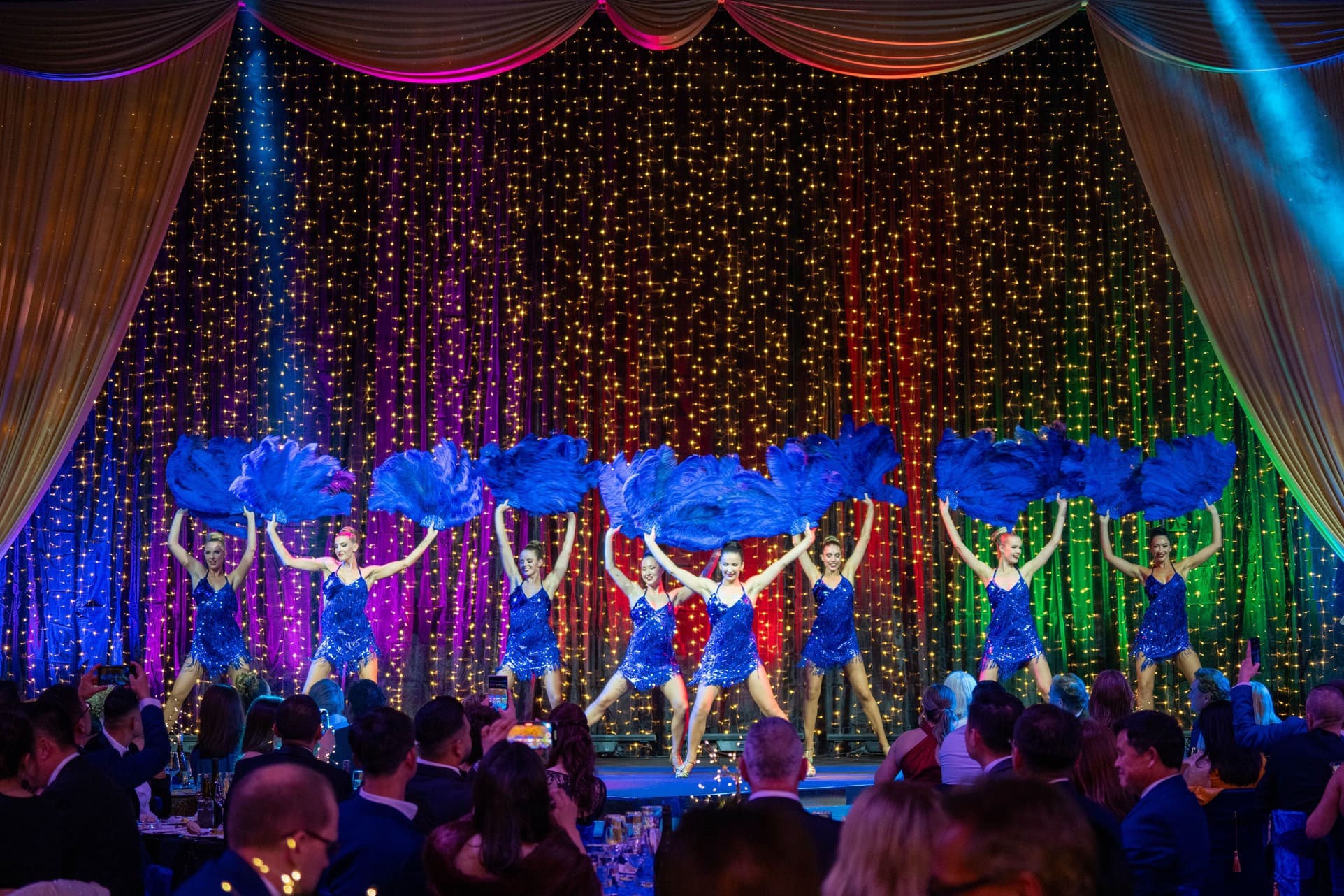 A group of dancers in blue dresses perform on stage, each holding up large blue feather fans. Behind them, golden string lights and rainbow-colored curtains create a festive backdrop as an audience watches.