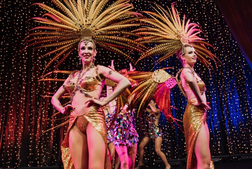 Two dancers in gold, sequined costumes and large feathered headdresses perform on stage with twinkling lights in the background. Other dancers in colorful costumes are visible behind them.