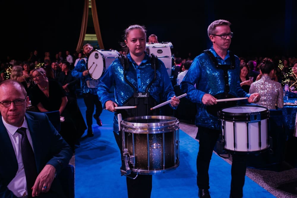 Two drummers in blue sequined vests perform while walking down a blue carpet at an indoor event, with seated guests and decorated tables in the background.