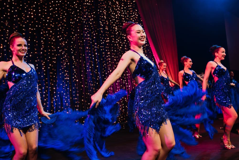 Five dancers in blue sequined dresses perform on stage, smiling and holding blue feathered props, with sparkling lights and red curtains in the background.