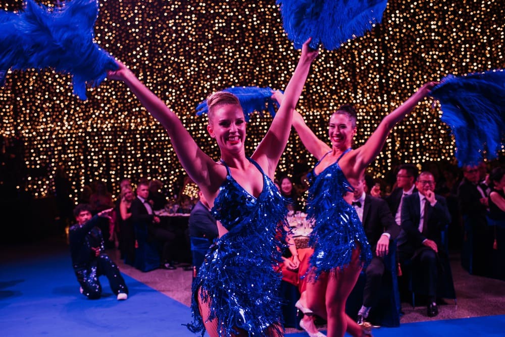 Two women in sparkling blue costumes dance with large blue feather fans in front of an audience. Warm lights twinkle in the background, creating a festive atmosphere. People in formal attire watch and smile.