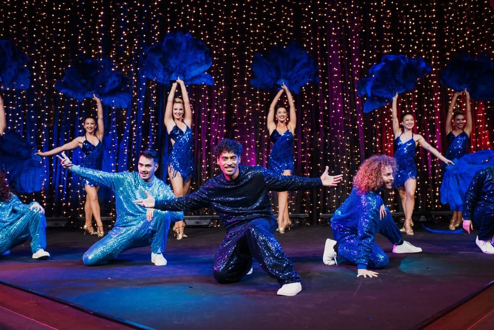 A group of dancers in blue sparkly costumes perform on stage, with women holding large blue feather fans in the background and three men posing energetically at the front. The backdrop is decorated with string lights.