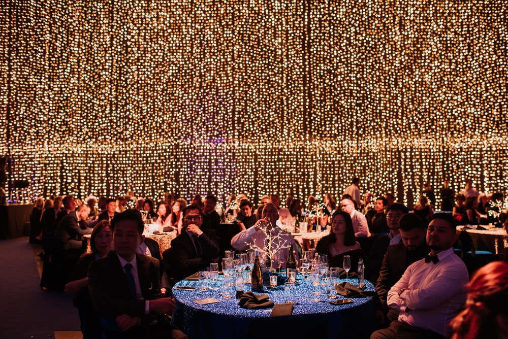 A large group of people dressed formally sit at round tables covered with blue cloth, surrounded by hundreds of warm, hanging lights creating a festive, elegant atmosphere.